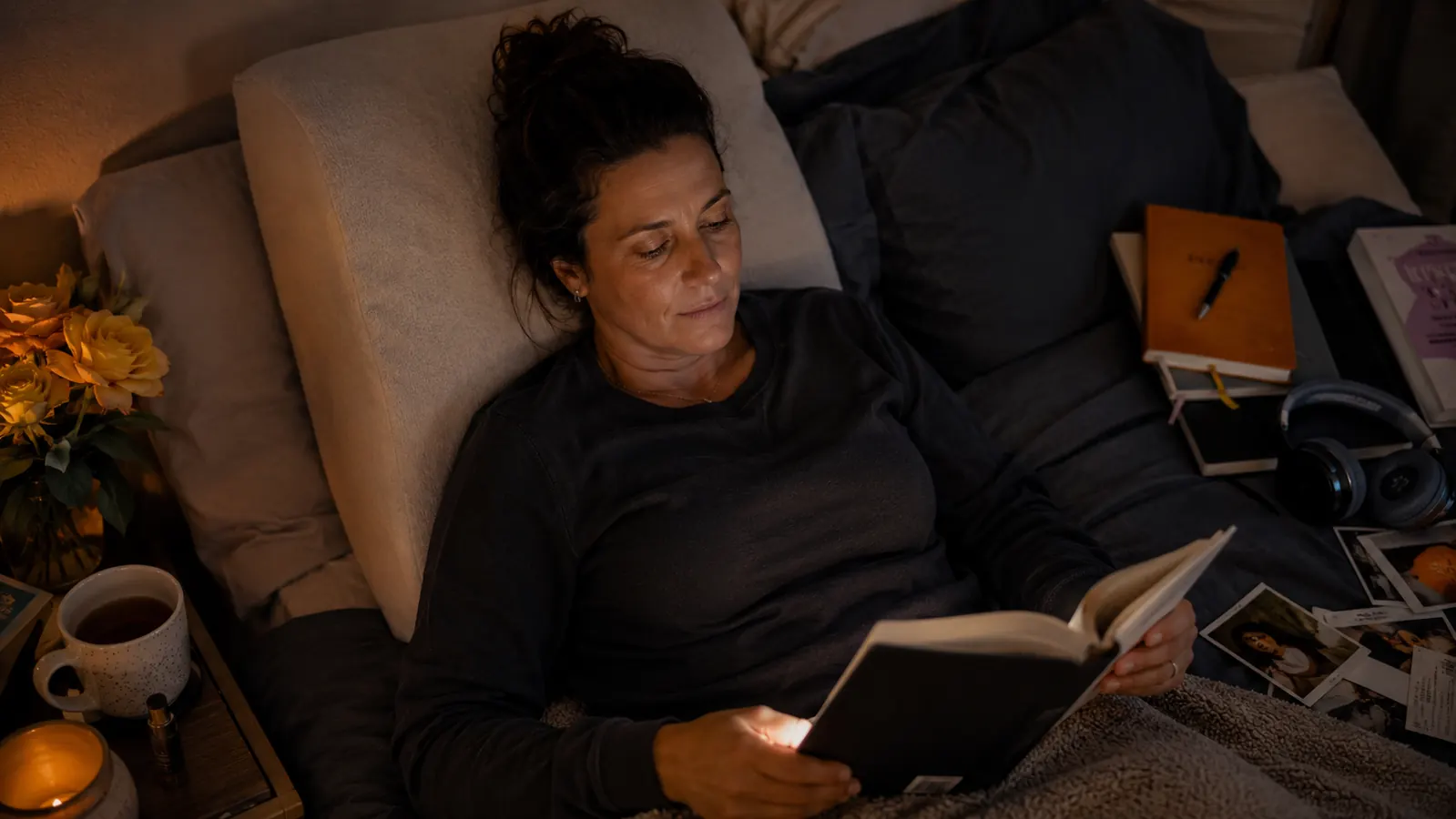 Person reclining against a reading pillow with arms, reading a paperback with a small clip-on light