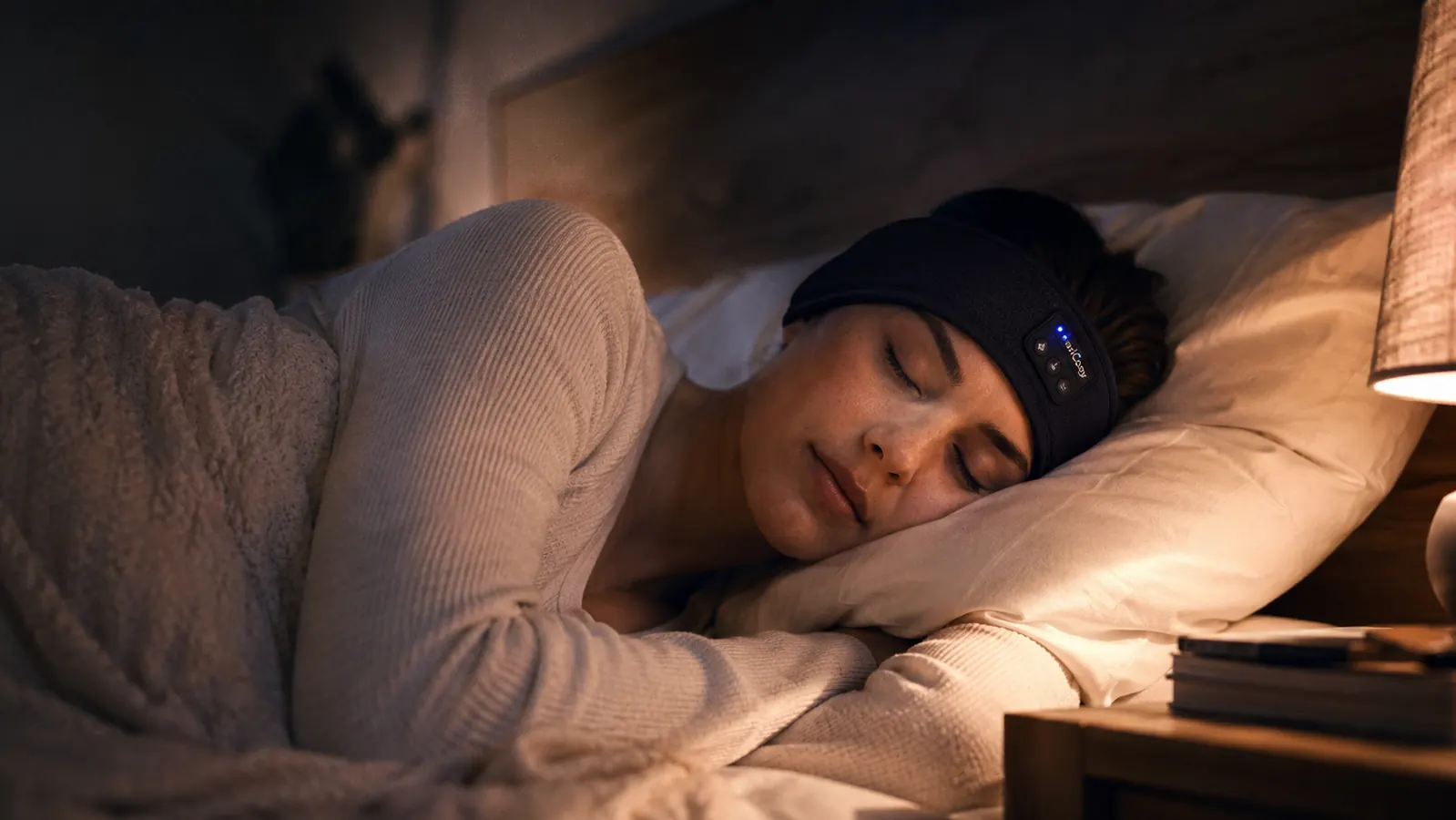 Woman lying on her side in bed at night wearing a thin fabric sleep headband, eyes closed, lamp glow in the background