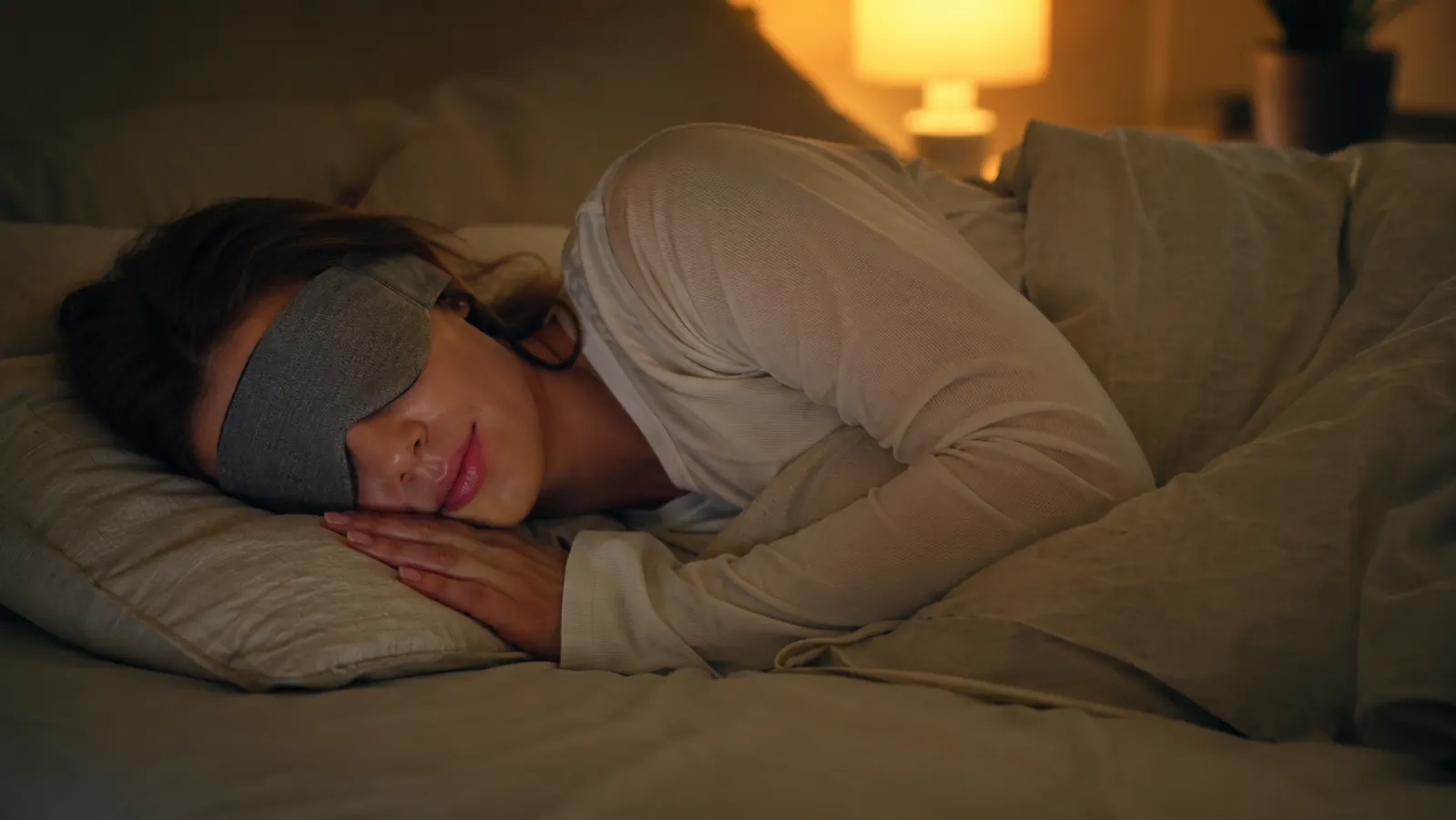 Woman lying on her side in bed at night wearing a soft fabric sleep headband, eyes closed, warm lamp glow in the background