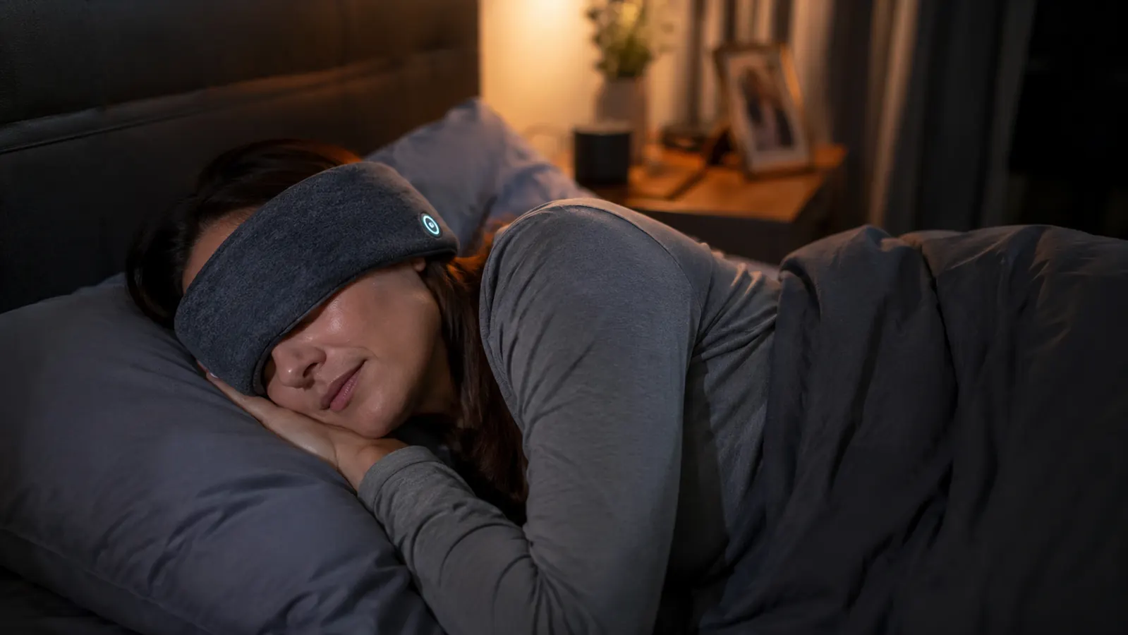 Woman lying on her side in bed at night wearing a thin fabric sleep headphone headband, room lit only by a dim bedside lamp