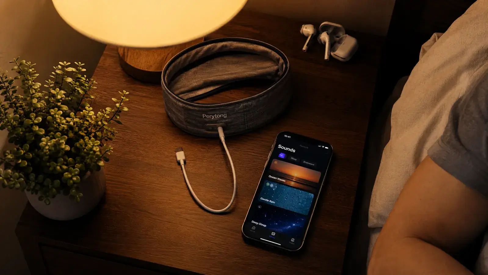Nightstand scene with the Perytong headband charging cable, a phone showing a sleep sounds app, and a folded set of earbuds pushed to the side