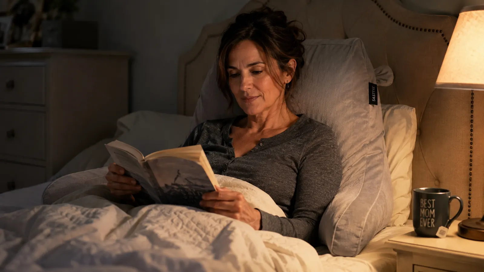 Woman sitting up in bed reading a paperback, back supported by a Sasttie reading pillow with armrests, warm bedside lamp light